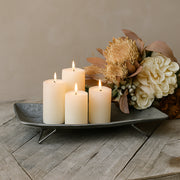 Rusztikus tálalási jelenet: bádog tál két gyertyával előtérben, mögötte fa tálcákon további gyertyák és szárított virágdekorációk láthatók.