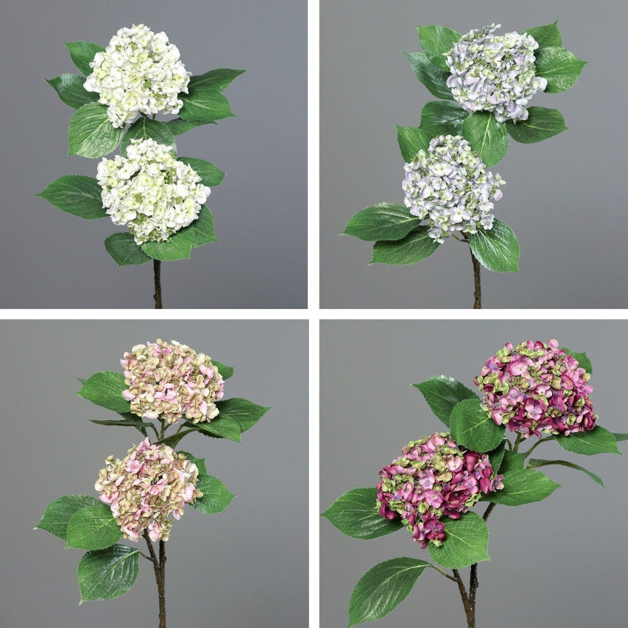 Mű hortenzia ág négyféle színváltozatban: fehér, halványlila, halványpink és lila árnyalatú virágfejekkel, zöld levelekkel, szürke háttér előtt.