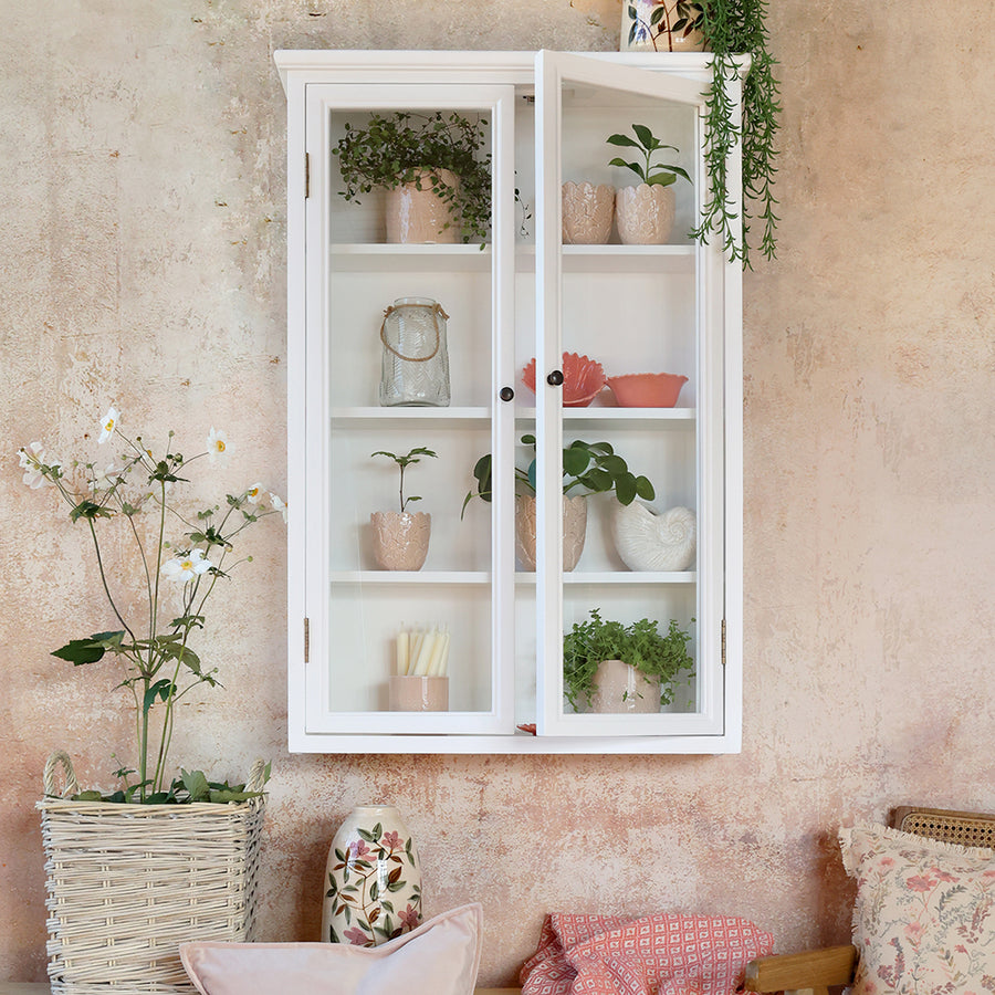 Old French fehér faliszekrény rusztikus falon, zöld növényekkel és rózsaszín kerámiákkal dekorálva.