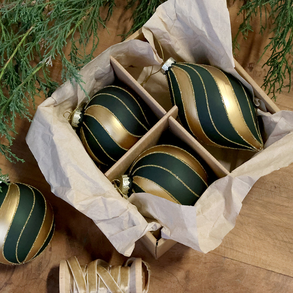 Chic Antique olívazöld-arany üvegdíszek papírral bélelt dobozban, fa felületen, örökzöld ágak és aranyszínű szalag mellett.