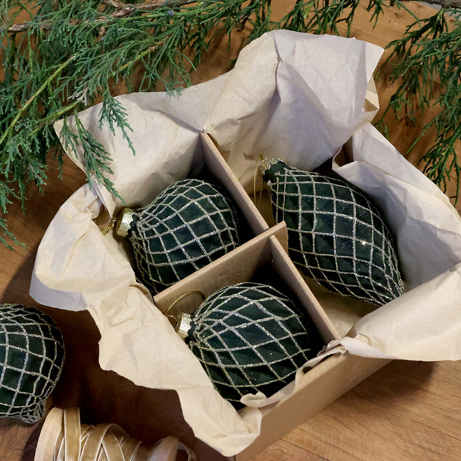 Chic Antique olívazöld hálómintás üvegdíszek papírral bélelt dobozban, fa felületen, örökzöld ágak mellett.