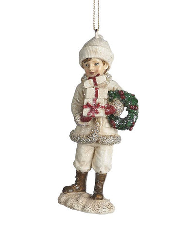 Ajándékot és ünnepi koszorút tartó, krémszínű téli kisfiú formájú karácsonyfadísz.