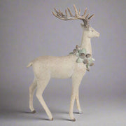 Tobozkoszorúval díszített, bézs színű, karácsonyi szarvas figura.