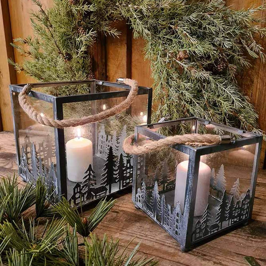 Vidéki rusztikus stílusú, antikolt szürke színű 2 darabos karácsonyi sziluettes fém lámpásszett fenyőkoszorú előtt antikolt fa deszkaasztalon 