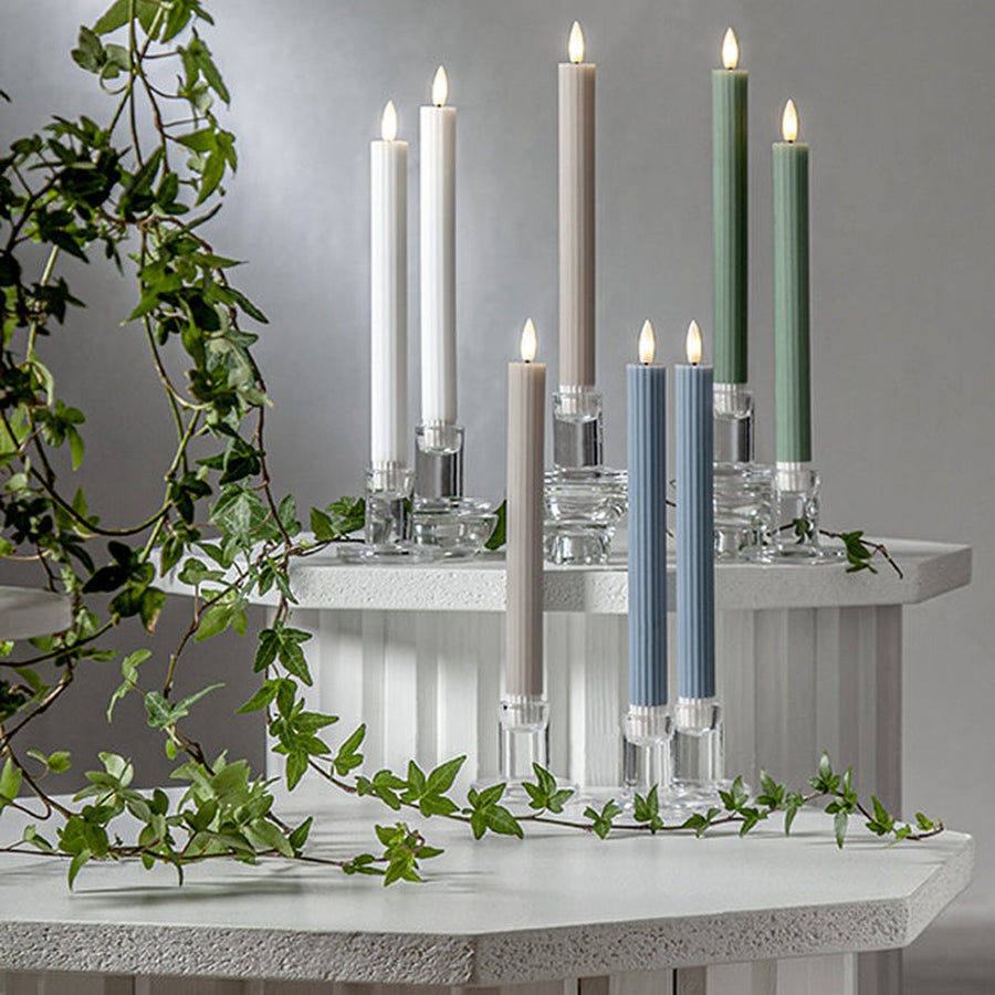 Pislákoló fényű LED vacsoragyertyák poharakban, hangulatos, természetes környezetben, zöld ágakkal körülvéve.