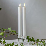 Fehér LED vacsoragyertyák dekor pohárban, meleg fényű lánggal, természetes zöld díszítéssel és elegáns hangulatban elrendezve.