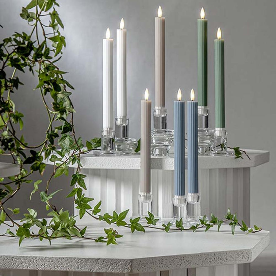 Pislákoló fényű LED vacsoragyertyák poharakban, hangulatos, természetes környezetben, zöld ágakkal körülvéve.