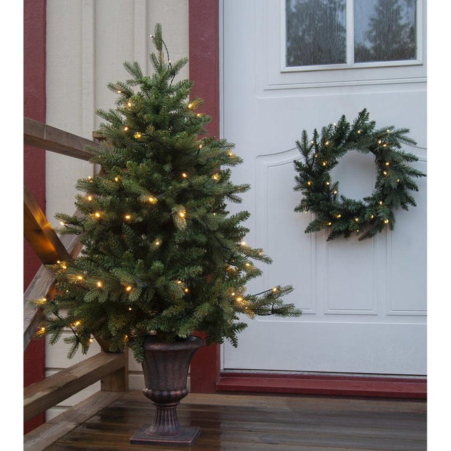 Élethű műfenyő, 3D ágakkal, beépített LED világítással, talpas kaspóban bejárati ajtó előtt.