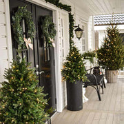 Élethű műfenyő, 3D ágakkal, beépített LED világítással, talpas kaspóban bejárati ajtó előtt.