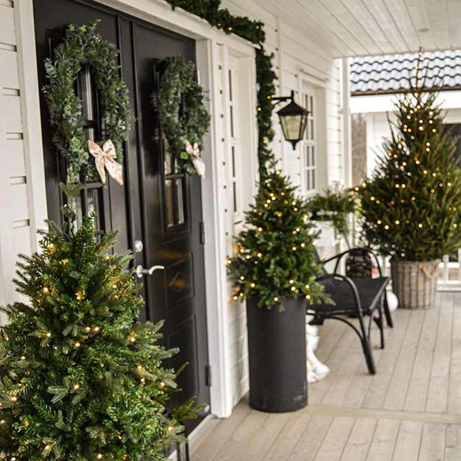 Élethű műfenyő, 3D ágakkal, beépített LED világítással, talpas kaspóban bejárati ajtó előtt.