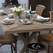 X-lábú, fenyőfából készült étkezőasztal fényfős ebédhez terítve.