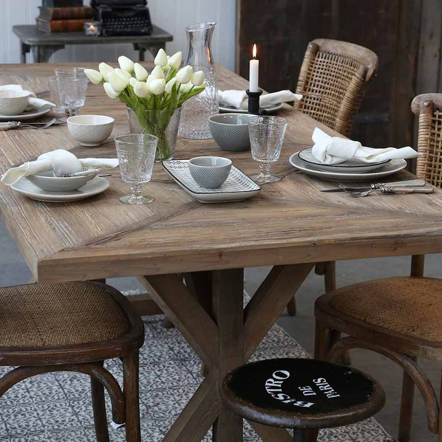 X-lábú, fenyőfából készült étkezőasztal fényfős ebédhez terítve.
