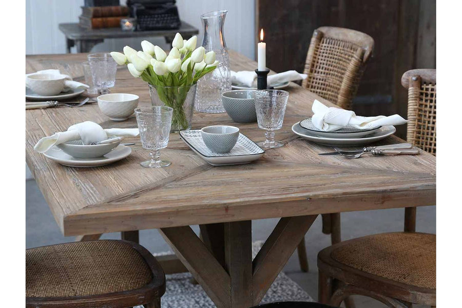 X-lábú, fenyőfából készült étkezőasztal fényfős ebédhez terítve.