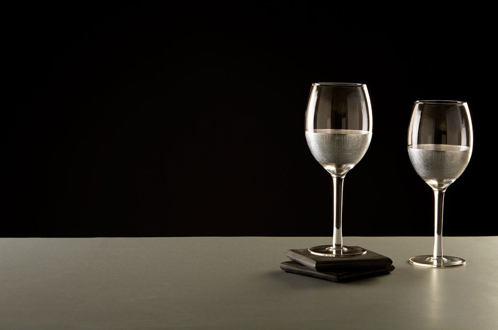 Modern, ezüstszínű fémmel díszített, üveg fehérboros poharak szürke asztalon, fekete háttér előtt.