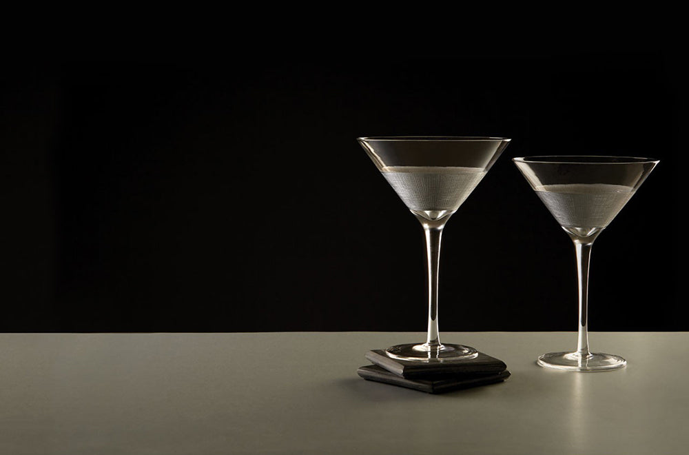 Modern, ezüstszínű fémmel díszített, üveg koktélospoharak szürke asztalon, fekete háttér előtt.