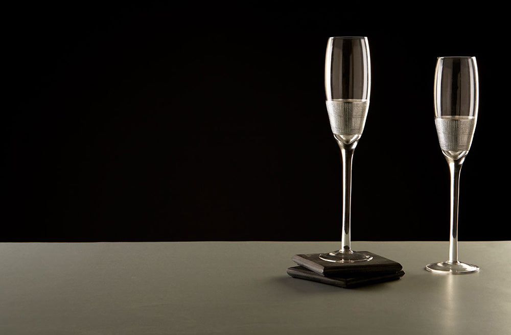 Modern, ezüstszínű fémmel díszített, üveg  pezsgőspoharak szürke asztalon, fekete háttér előtt.