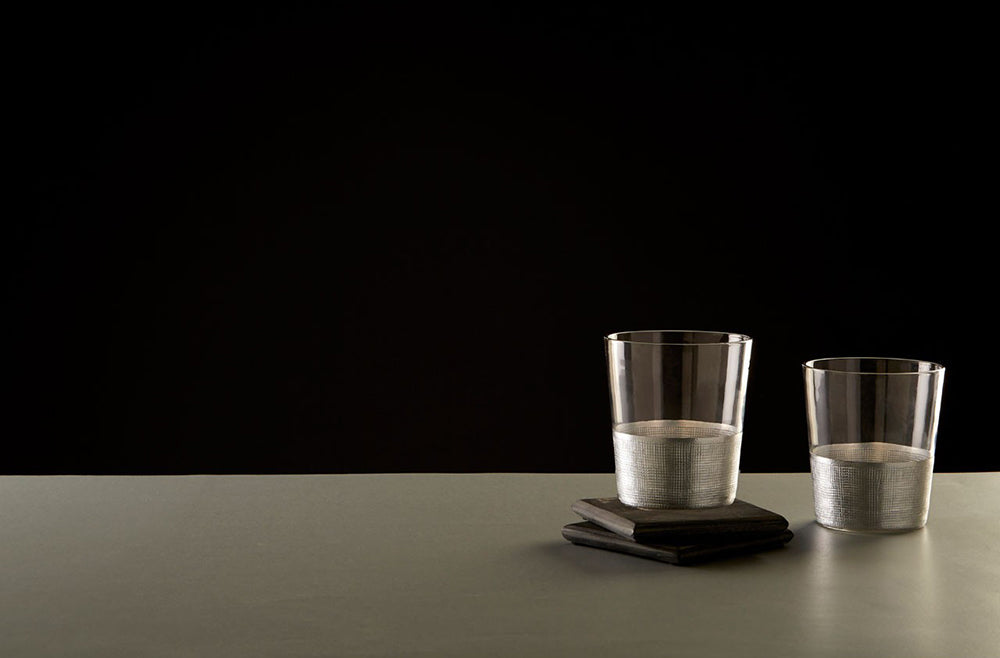Modern, ezüstszínű fémmel díszített, röviditalos üveg poharak szürke asztalon, fekete háttér előtt.