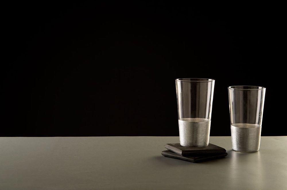 Modern, ezüstszínű fémmel díszített, üveg vizespoharak szürke asztalon, fekete háttér előtt.