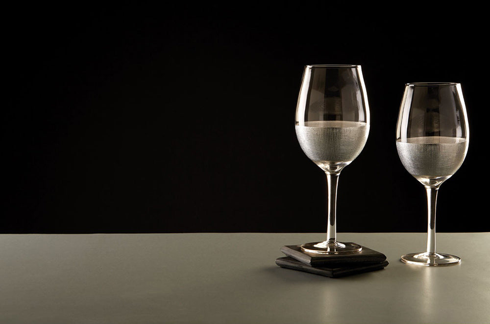 Modern, ezüstszínű fémmel díszített, üveg vörösboros poharak szürke asztalon, fekete háttér előtt.