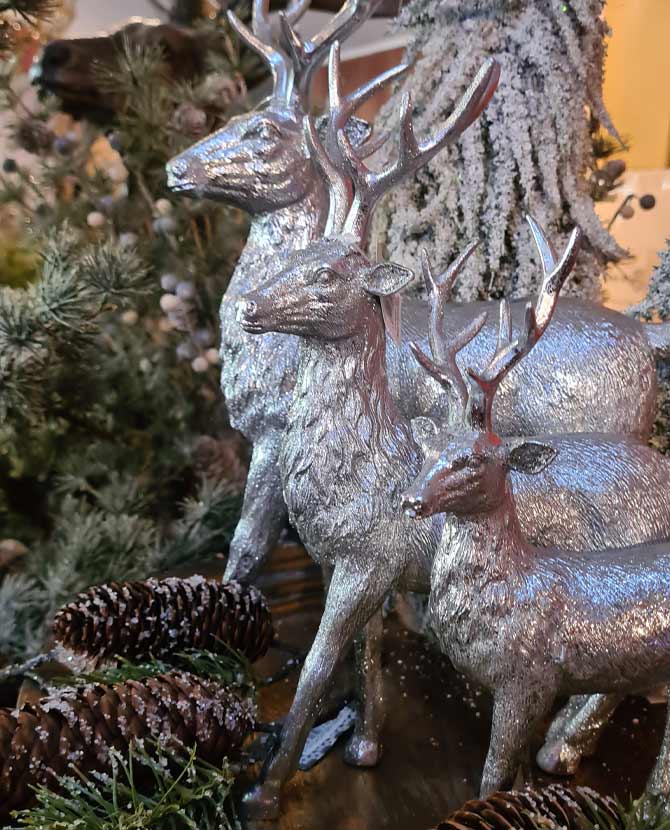 Glamour stílusú, 21 cm magas, ezüst színű karácsonyi szarvas figura 3 változatban fenyők és tobozok között