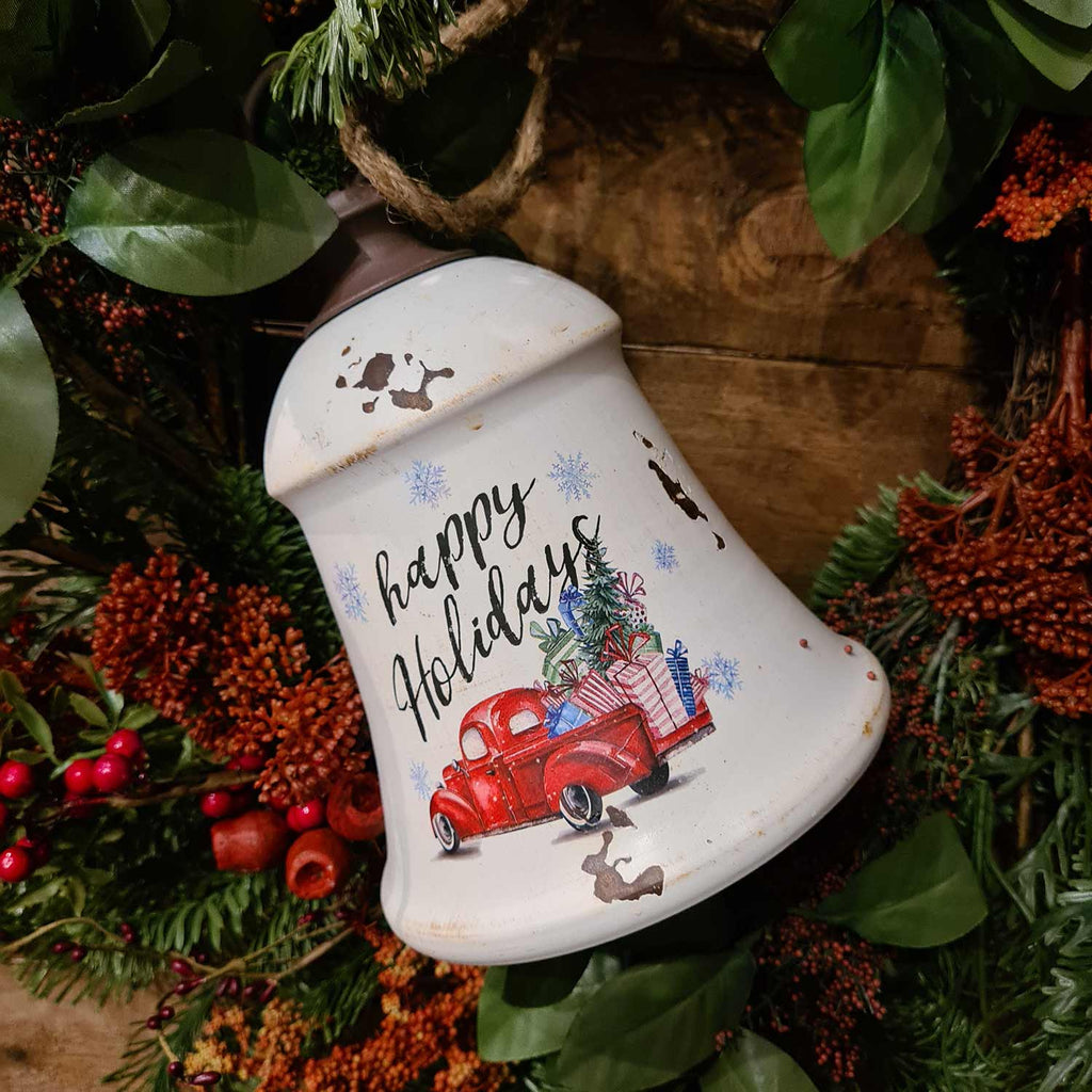 Vidéki, farmhouse stílusú antikolt, kopott fehér színű, Happy Holidays feliratú, ajándékokkal megrakott, piros színű platós farm kocsival díszített, fémből készült karácsonyi harang.