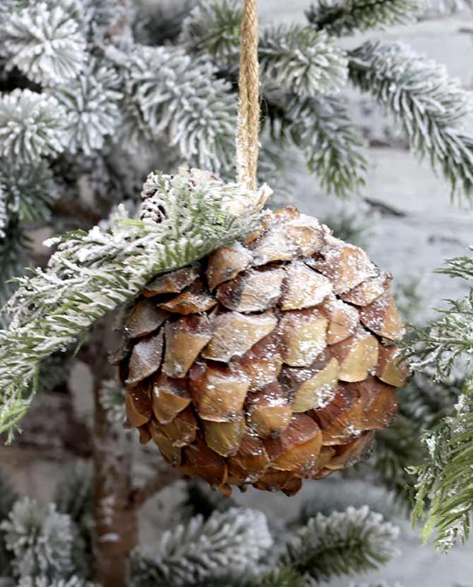 Vidéki stílusú, 12 cm átmérőjű, fenyőággal díszített, havas felületű, karácsonyi toboz gömb dísz, havas fenyőágon.