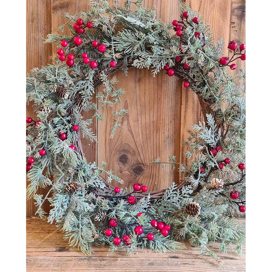 Piros bogyókkal díszített, ezüstös zöld színű mesterséges fenyőágakból készült karácsonyi koszorú, ajtódísz. fa fal előtt.