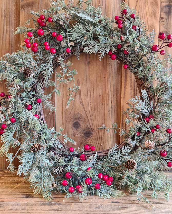 Piros bogyókkal díszített, ezüstös zöld színű mesterséges fenyőágakból készült karácsonyi koszorú, ajtódísz. fa fal előtt.