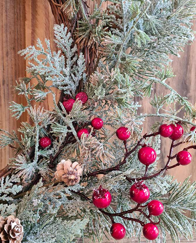 Piros bogyókkal díszített, ezüstös zöld színű mesterséges fenyőágakból készült karácsonyi koszorú, ajtódísz.