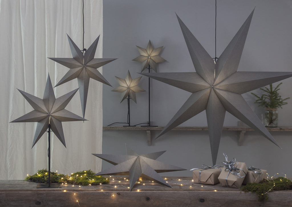 Különböző méretű, szürkeszínű karácsonyi papírcsillag lámpák, ajándékokkal és zöld fenyőágakkal.