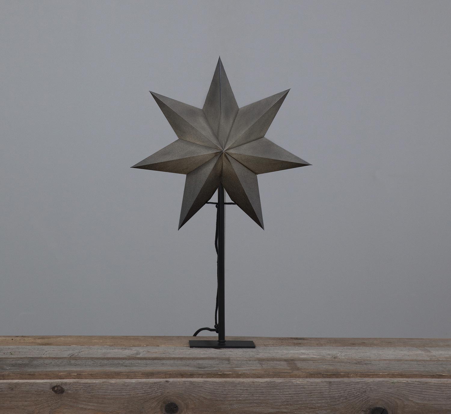 Szürkeszínű karácsonyi pípírcsillag fém talapzaton, natúr fa asztalon.
