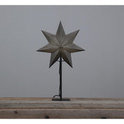 Szürkeszínű karácsonyi pípírcsillag fém talapzaton, natúr fa asztalon.