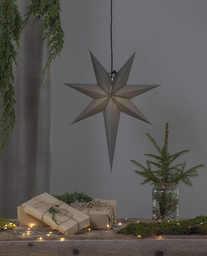 Szürkeszínű, karácsonyi papírcsillag lámpa szürke háttér előtt, fenyőágakkal.