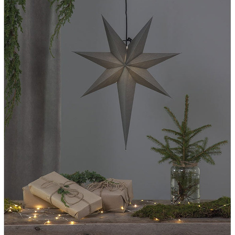 Szürkeszínű, karácsonyi papírcsillag lámpa szürke háttér előtt, fenyőágakkal.