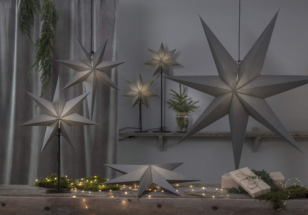 Különböző méretű, szürkeszínű karácsonyi papírcsillag lámpák, ajándékokkal és zöld fenyőágakkal..