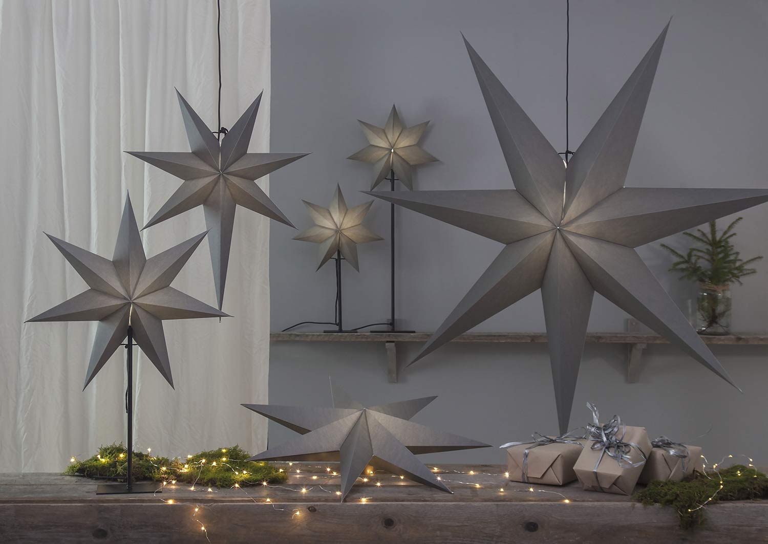 Különböző méretű, szürkeszínű karácsonyi papírcsillag lámpák, ajándékokkal és zöld fenyőágakkal..