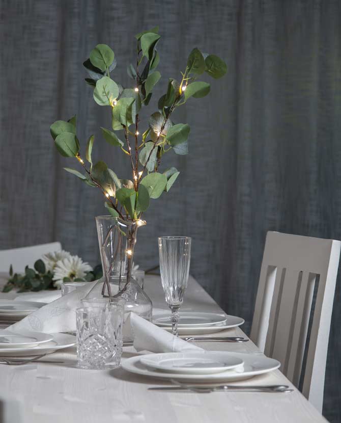 Élethű, 90 cm magas, mesterséges eukaliptusz ág műnövény, beépített, időzíthető meleg fehér fényű, harmatcsepp formájú LED világítással, ünnepi fehér asztalon, szürke háttér előtt , üveg borospoharakkal és  fehér színű terítékkel 