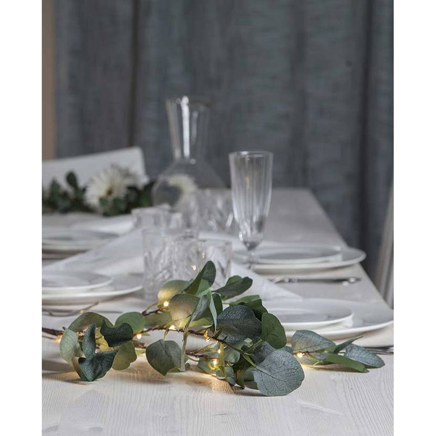 Élethű, 90 cm magas, mesterséges eukaliptusz ág műnövény, beépített, időzíthető meleg fehér fényű, harmatcsepp formájú LED világítással, ünnepi fehér asztalon, szürke háttér előtt , üveg borospoharakkal és fehér színű terítékkel