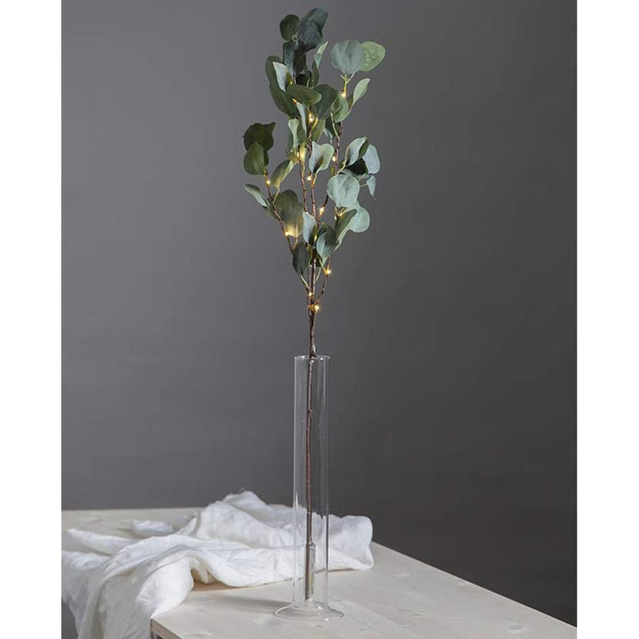 Led-es mesterséges eukaliptusz ág műnövény, henger alakú üvegvázában , fehér asztalon, szürke fal előtt 