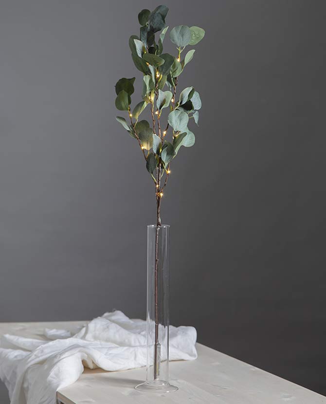 Led-es mesterséges eukaliptusz ág műnövény, henger alakú üvegvázában , fehér asztalon, szürke fal előtt 