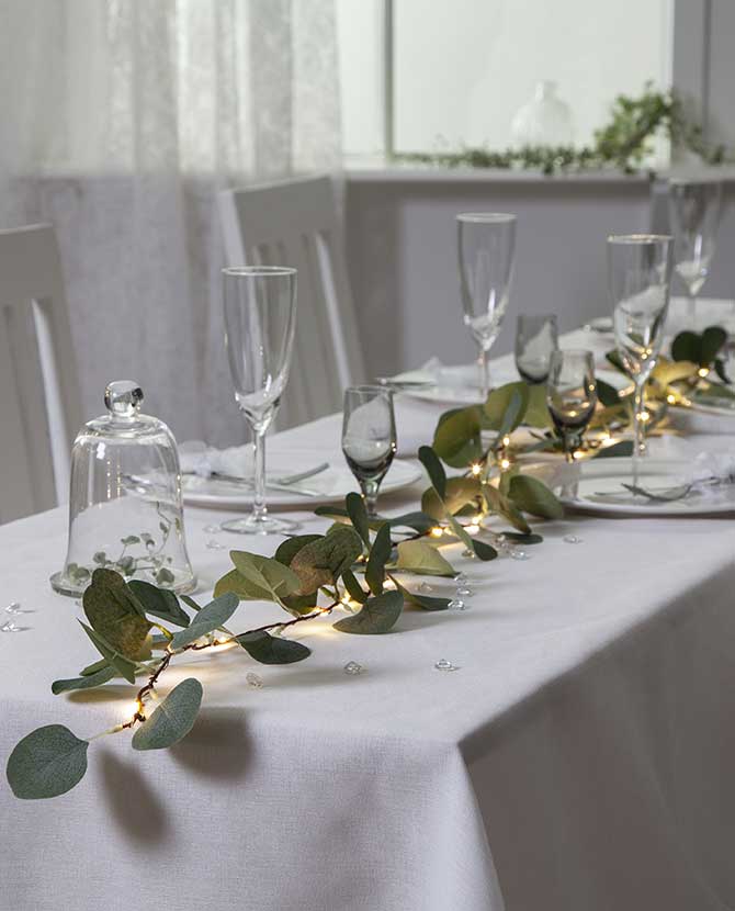 Élethű, 180 cm hosszú, mesterséges eukaliptusz girland műnövény, beépített, időzíthető meleg fehér fényű, harmatcsepp formájú LED világítással , ünnepi, fehér asztalterítőjű asztalon, borospoharakkal és fehér étkészlettel. 