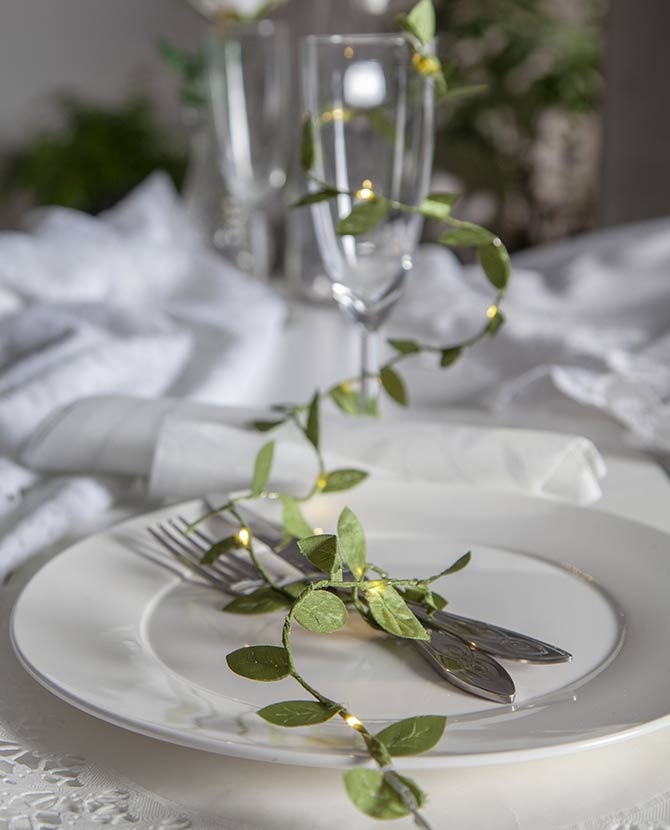 Mesterséges, 95 cm hosszú apró, zöld levelű girland műnövény, meleg fehér fényű, harmatcsepp formájú LED világítással ünnepi fehér terített asztalon