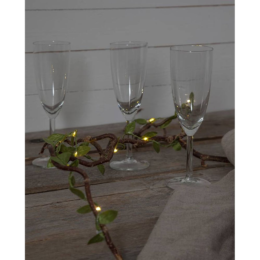 Mesterséges, 95 cm hosszú apró, zöld levelű girland műnövény, meleg fehér fényű, harmatcsepp formájú LED világítással barna ágak között pezsgőspoharakkal
