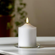 Valódi viaszból készült, fehér színű led gyertya üveg tányéron, fehér színű asztalon, szürke fal előtt