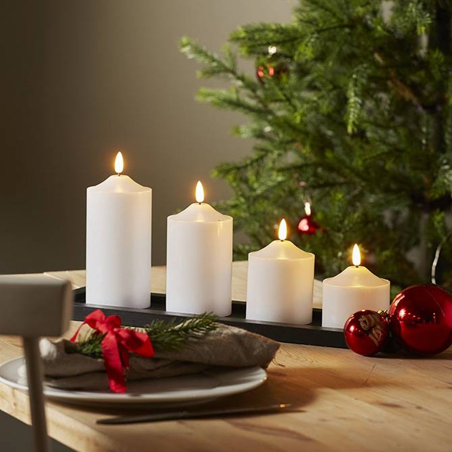 Karácsonyi hangulatú, fehér színű led gyertyák. 4 féle méretben szürke színű tálcán, fa asztalon, háttérben fenyőfával