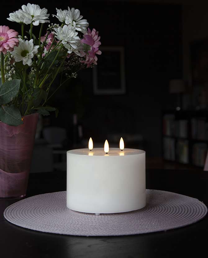 Fehér színű, viaszból készült, 3 darab meleg fehér fényű led lángú led gyertya fekete asztalon, lila és fehér színű virágokkal sötét szobában