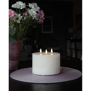 Fehér színű, viaszból készült, 3 darab meleg fehér fényű led lángú led gyertya fekete asztalon, lila és fehér színű virágokkal sötét szobában