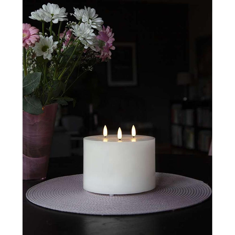 Fehér színű, viaszból készült, 3 darab meleg fehér fényű led lángú led gyertya fekete asztalon, lila és fehér színű virágokkal sötét szobában