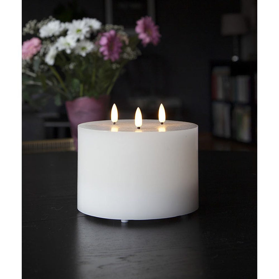 Fehér színű, viaszból készült, 3 darab meleg fehér fényű led lángú led gyertya fekete asztalon, lila és fehér színű virágokkal sötét szobában