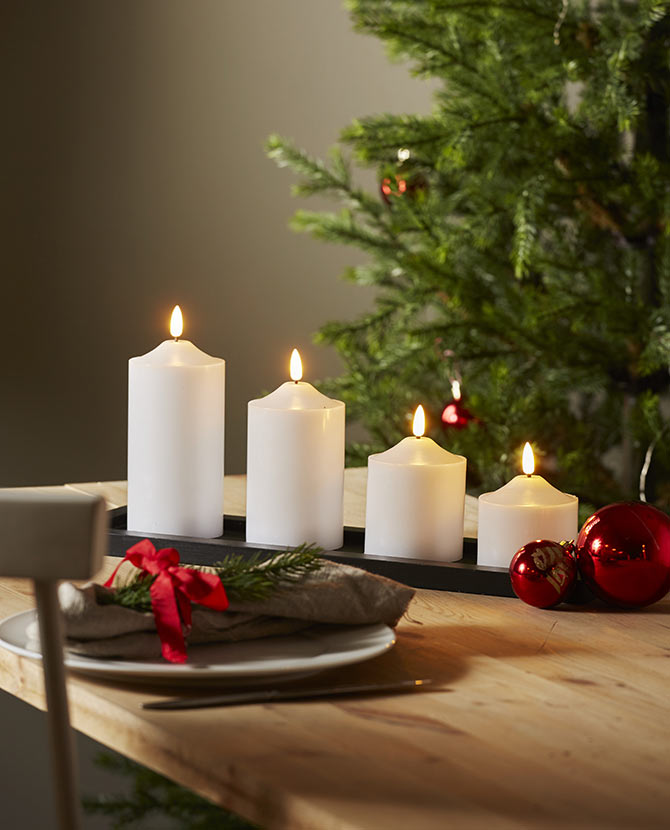 Karácsonyi hangulatú, fehér színű led gyertyák. 4 féle méretben szürke színű tálcán, fa asztalon, háttérben fenyőfával